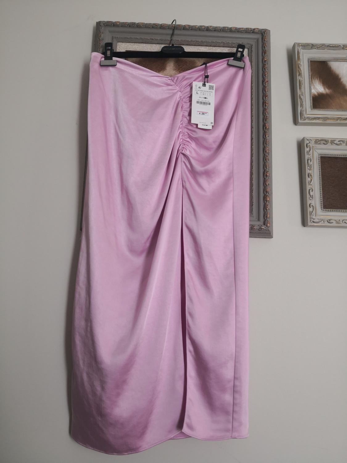 Light pink satin maxi skirt with central ruching, hanging on a black hanger in front of a decorative frame and beige wall.