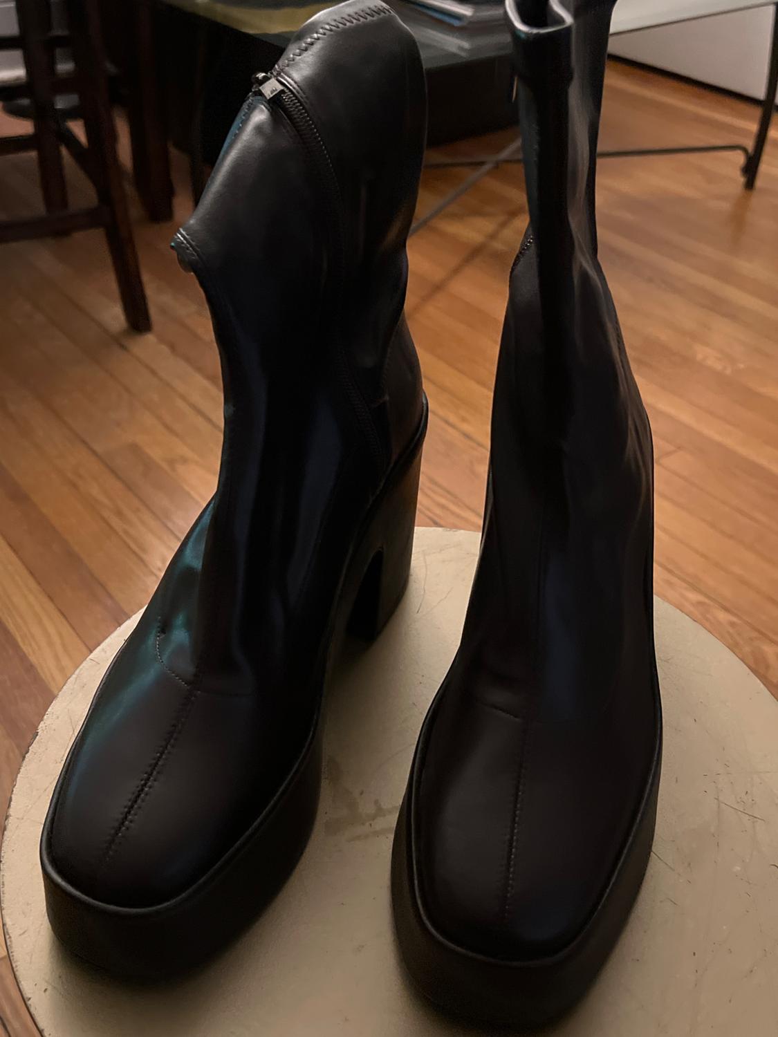 Black high-heeled boots with a side zip on a light wooden table in an indoor setting.