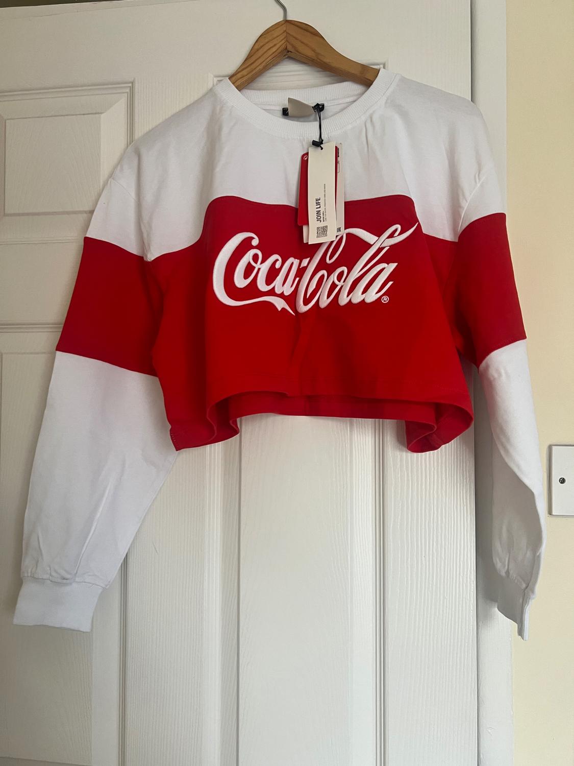White and red cropped sweatshirt with Coca-Cola logo on the chest, hanging on a wooden hanger over a white door.