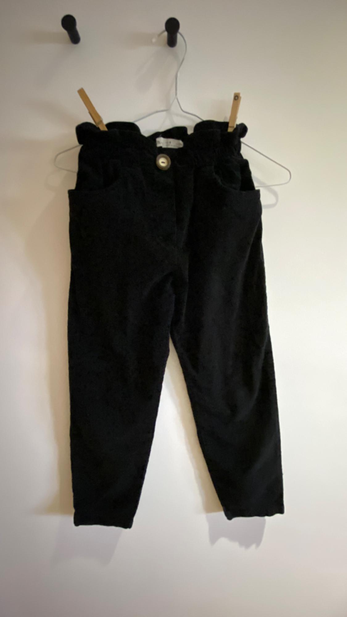 Black trousers hanging on a white hanger with wooden pegs, on a white wall with two black hooks.