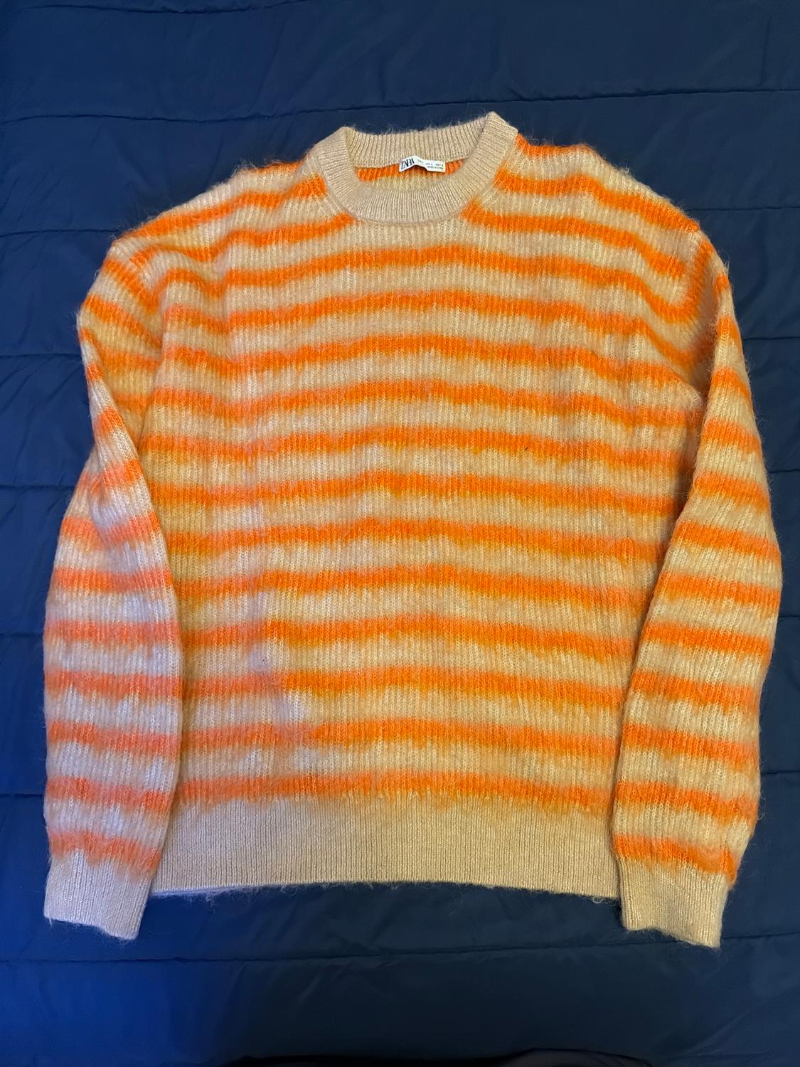 Horizontal striped jumper in shades of orange and beige on a dark blue background. Visible label on the collar.