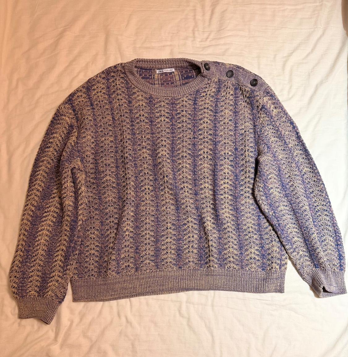 Knitted jumper in purple and beige tones with buttons on the left shoulder, on a light surface.