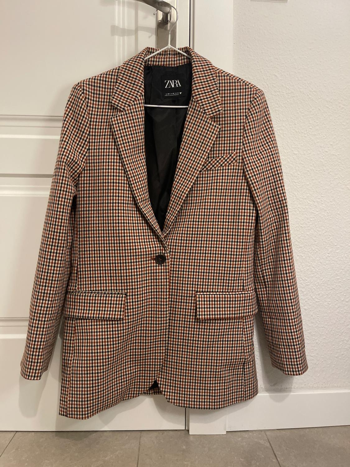 Brown and black checked jacket hanging on a hanger. Visible label with the text "ZARA". White wall and door background.