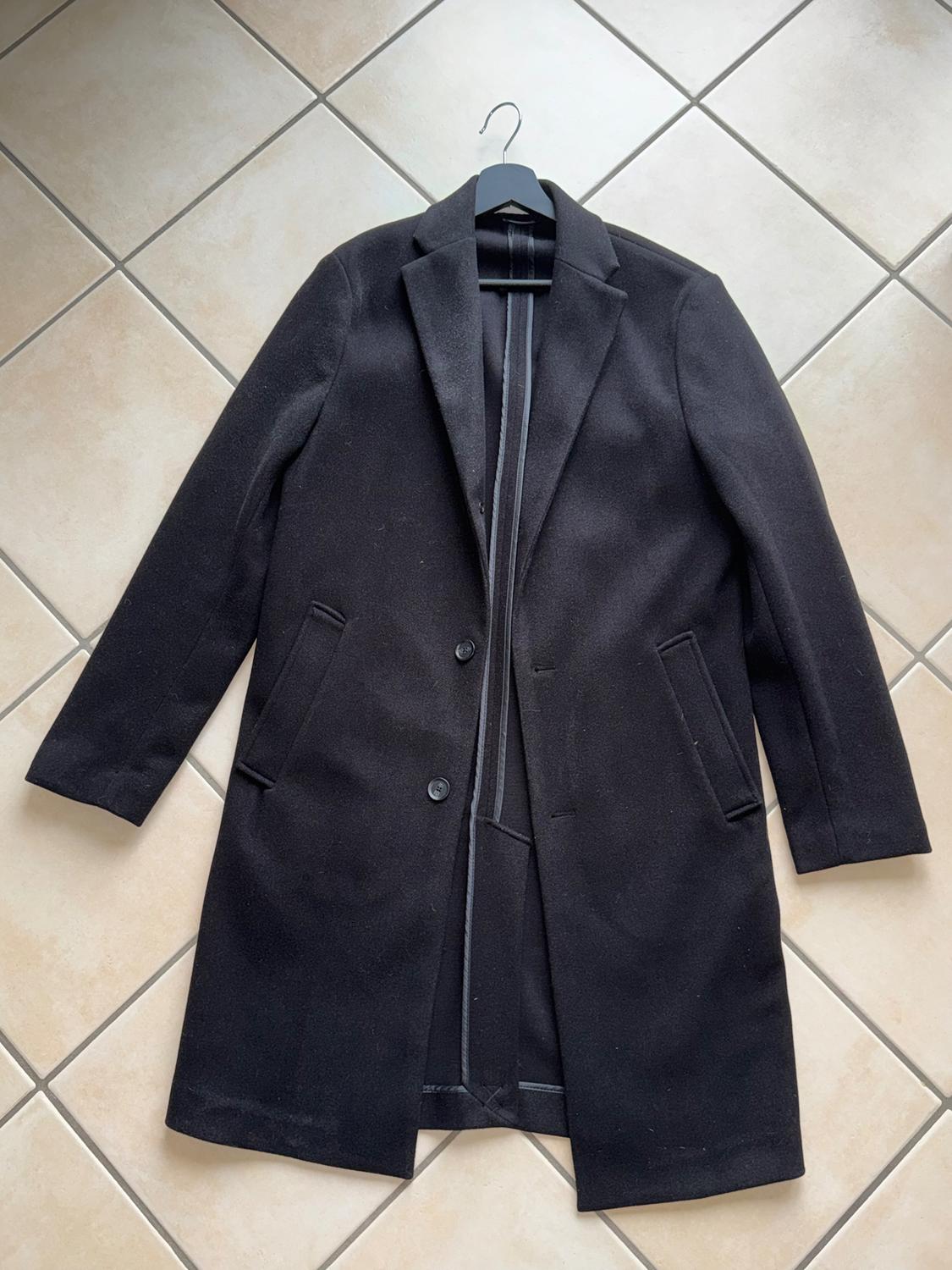 Black wool coat with lapels and two buttons, hanging on a hanger over a beige tiled floor.