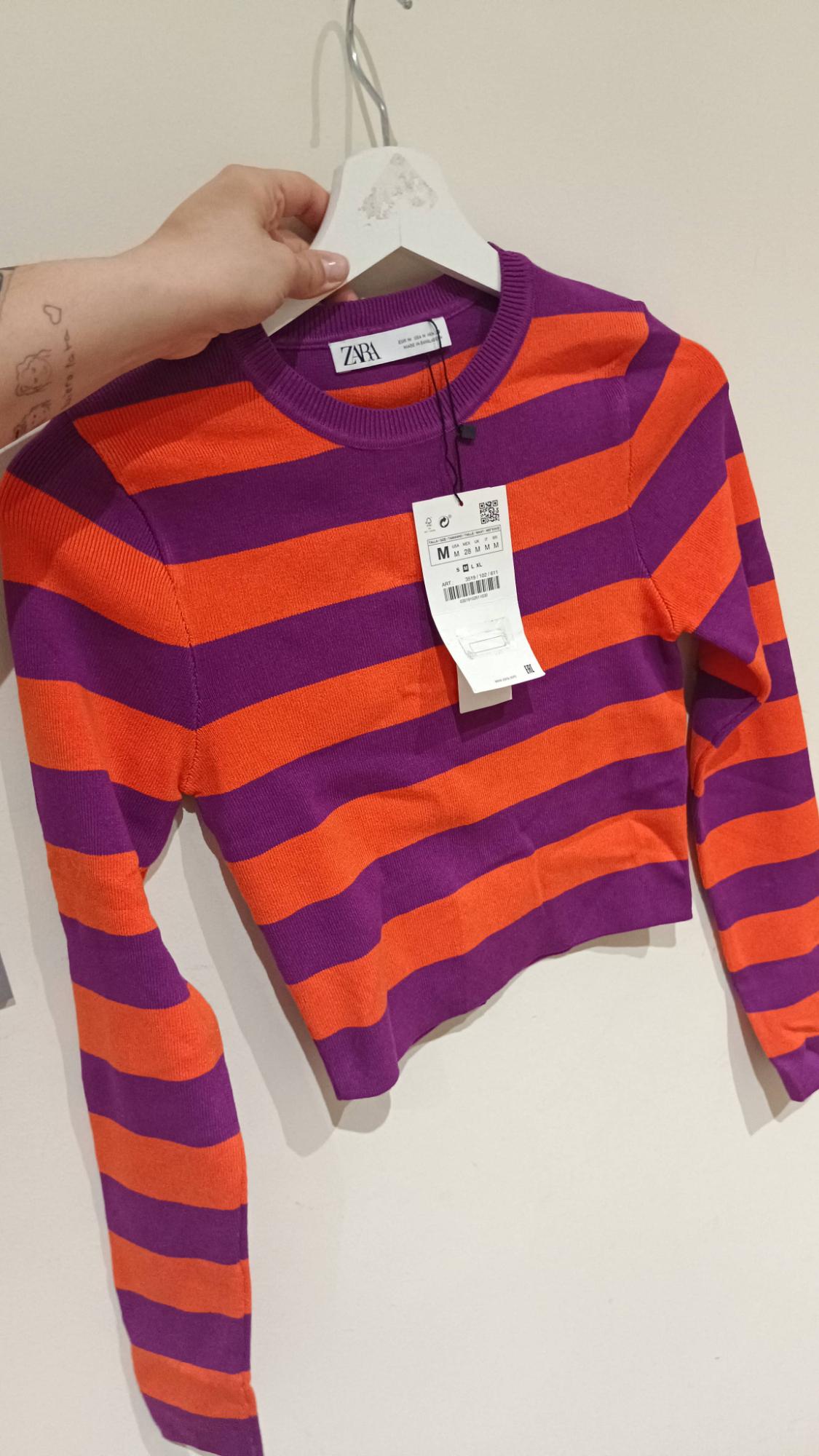 Orange and purple striped jumper hanging on a white hanger. Visible tag with size and price details. Hand holding the hanger.