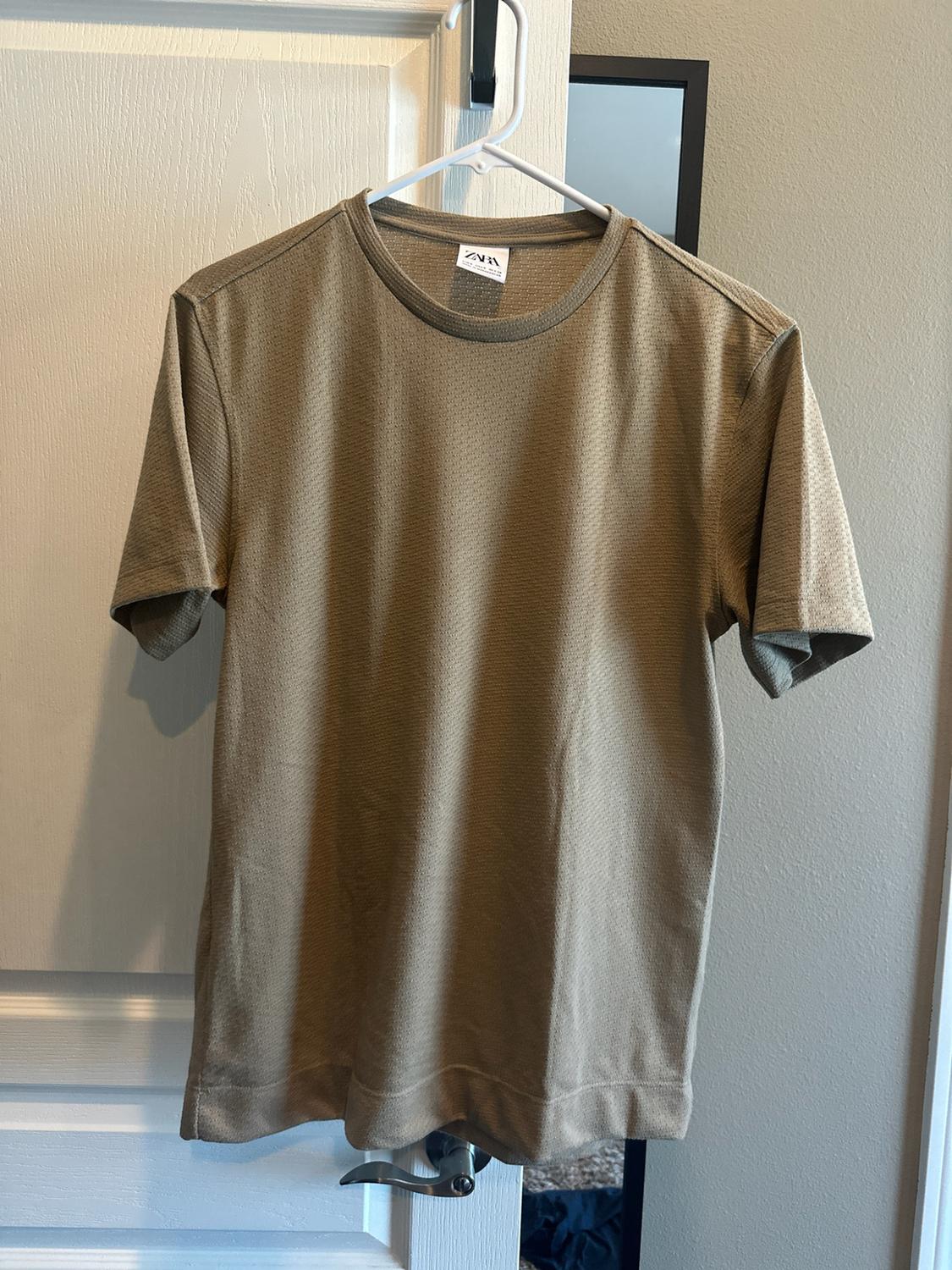 Textured short-sleeved sand-coloured T-shirt hanging on a white hanger in front of a door. Visible label bearing the text ZARA.