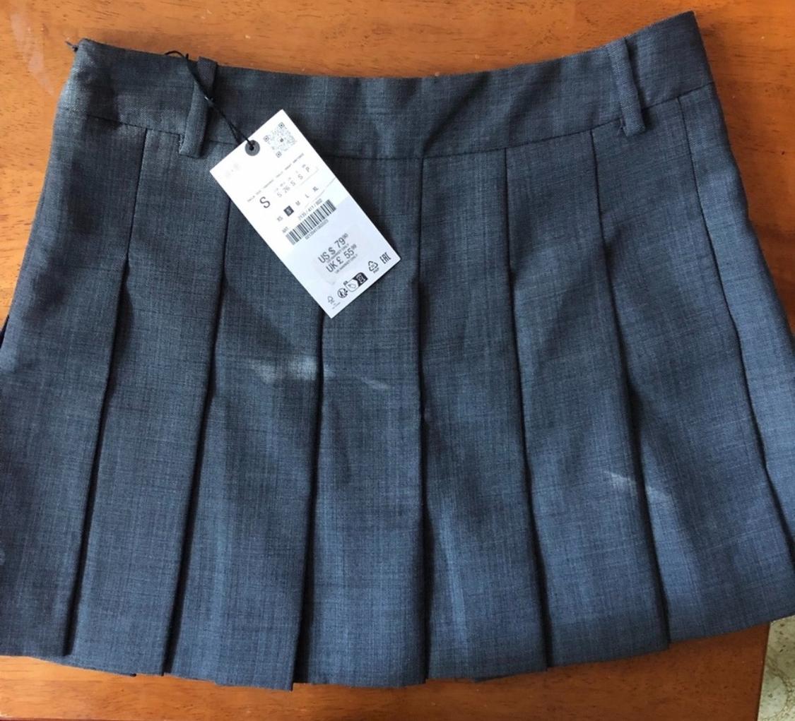 Gray pleated skirt on a wooden table, with a hanging tag showing size S and price US$ 7.99, UK£ 5.99.