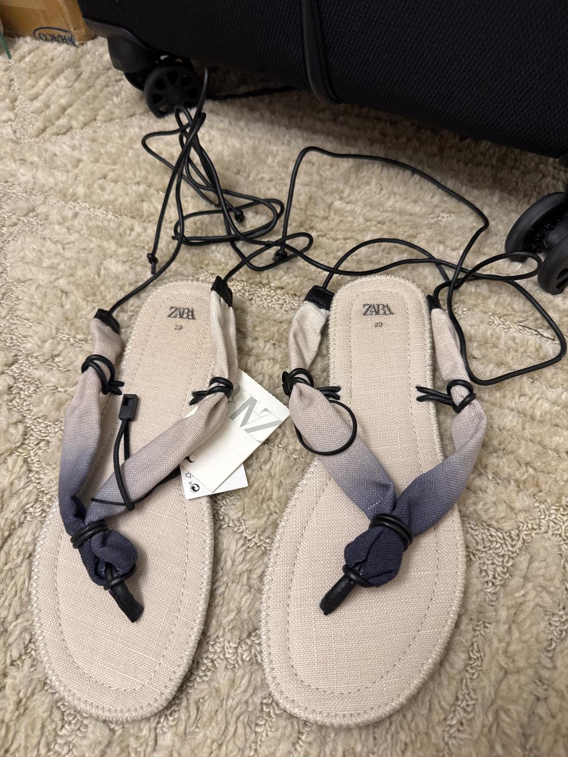 Beige fabric sandals with black and grey straps, on a beige carpet. Visible label with text "ZARA".