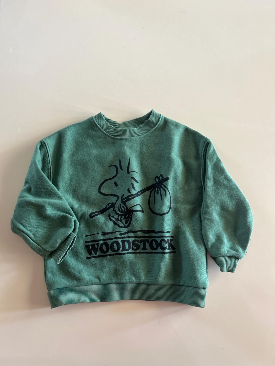 Green sweatshirt with a cartoon character design and the word "WOODSTOCK" in black, on a white background.