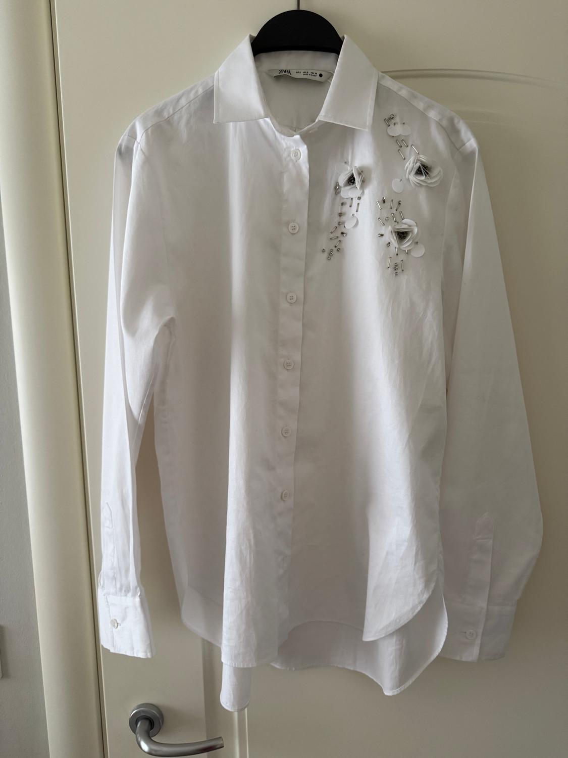 White shirt hanging on a door, with floral embroidery on the right shoulder.