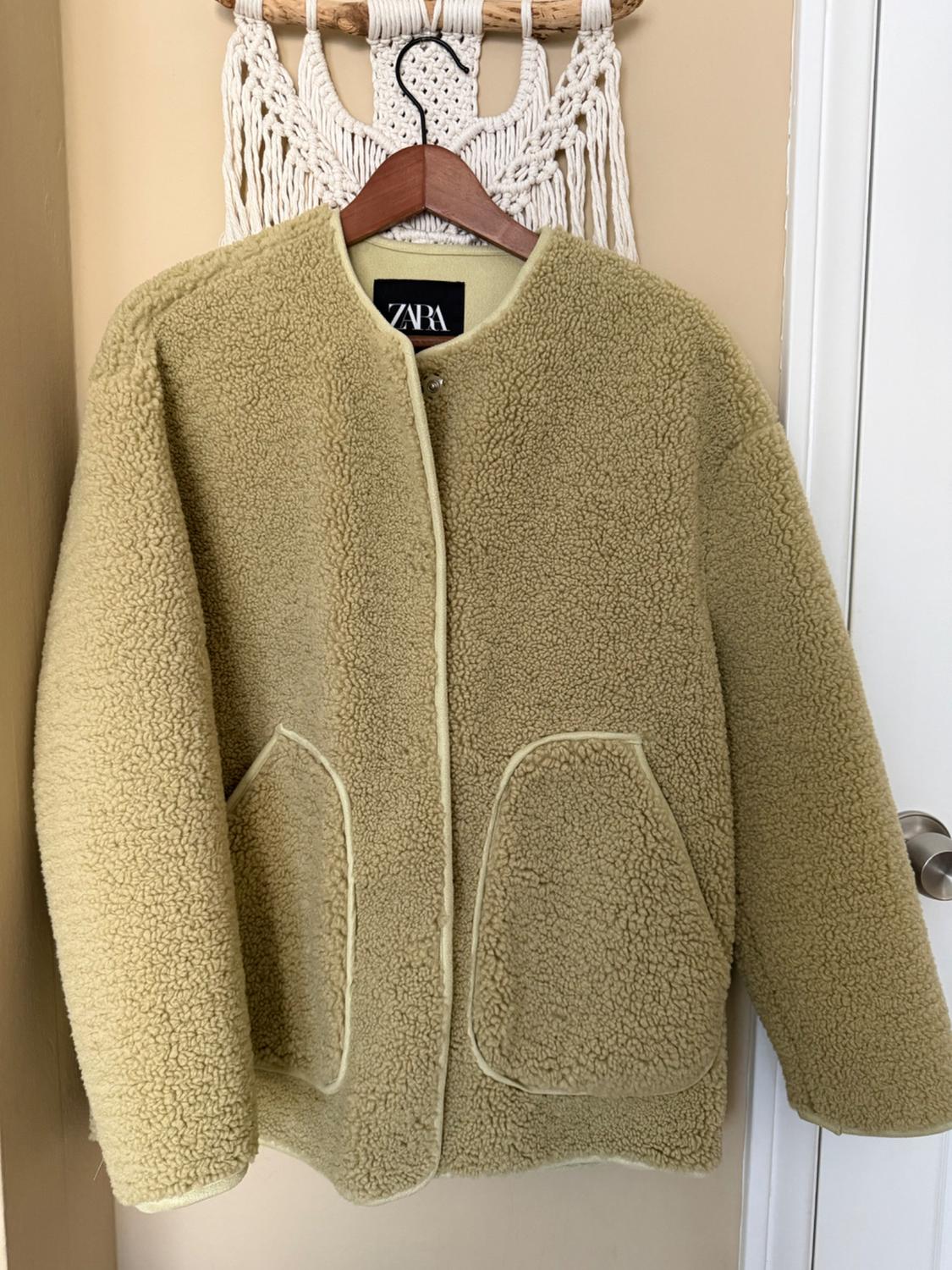 Beige wool jacket hanging on a wooden hanger with a "ZARA" tag, against a white macrame background and a beige wall.