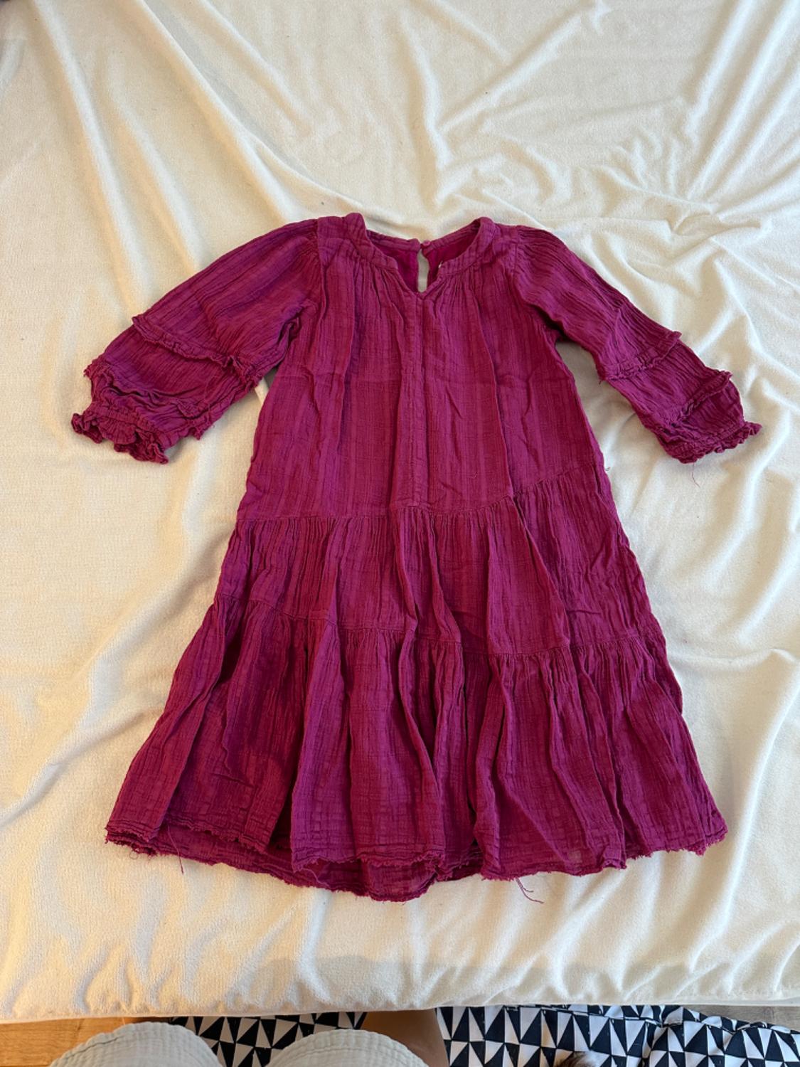 Purple, long-sleeved dress with frills, over wrinkled beige fabric. Geometric black and white background at the bottom.