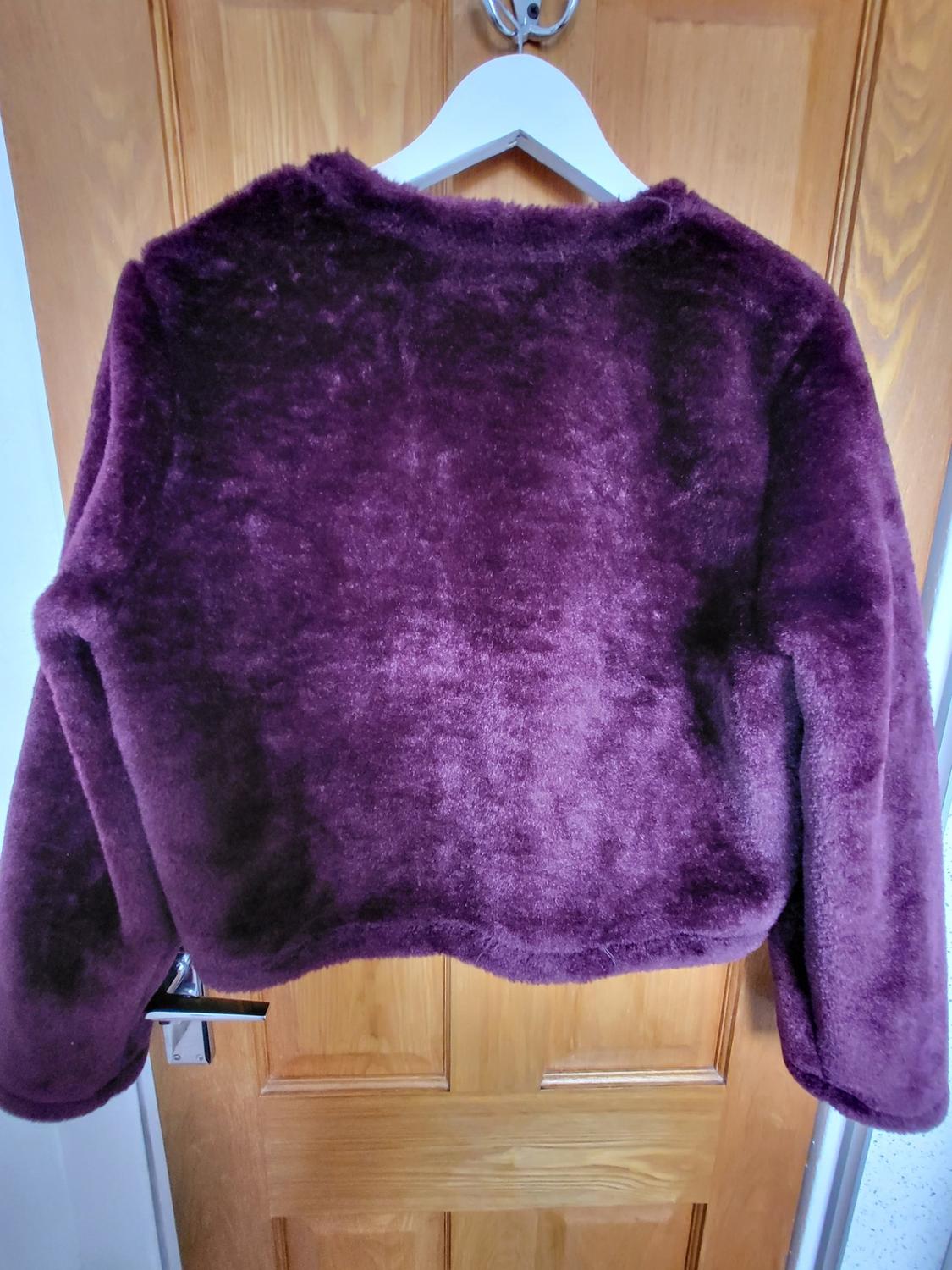 Short, faux-fur textured jacket in dark purple, hanging on a white hanger against a wooden door.
