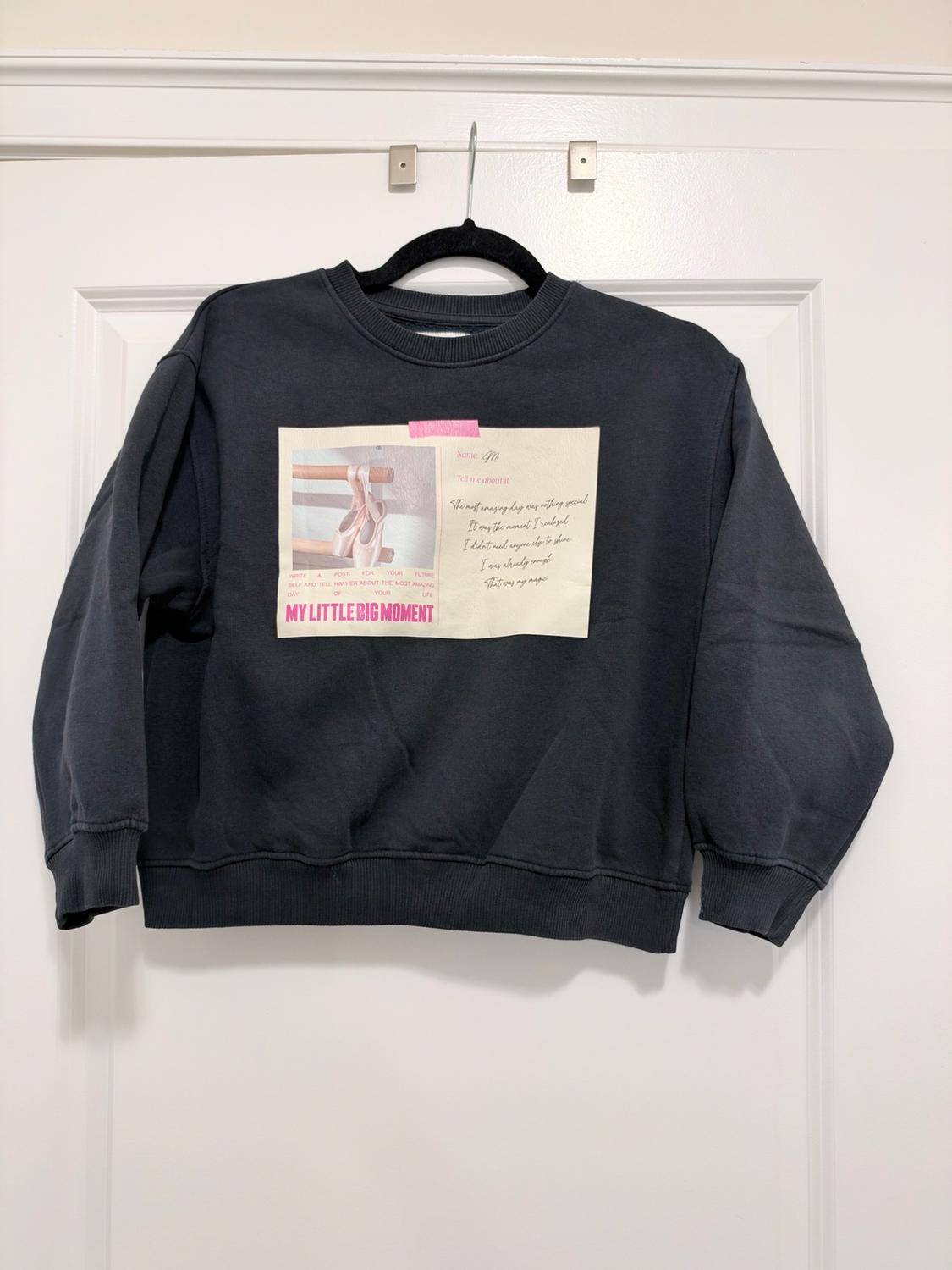 Dark gray sweatshirt hanging on a white door. It has a graphic
with ballet slippers and the text MY LITTLE BIG MOMENT.