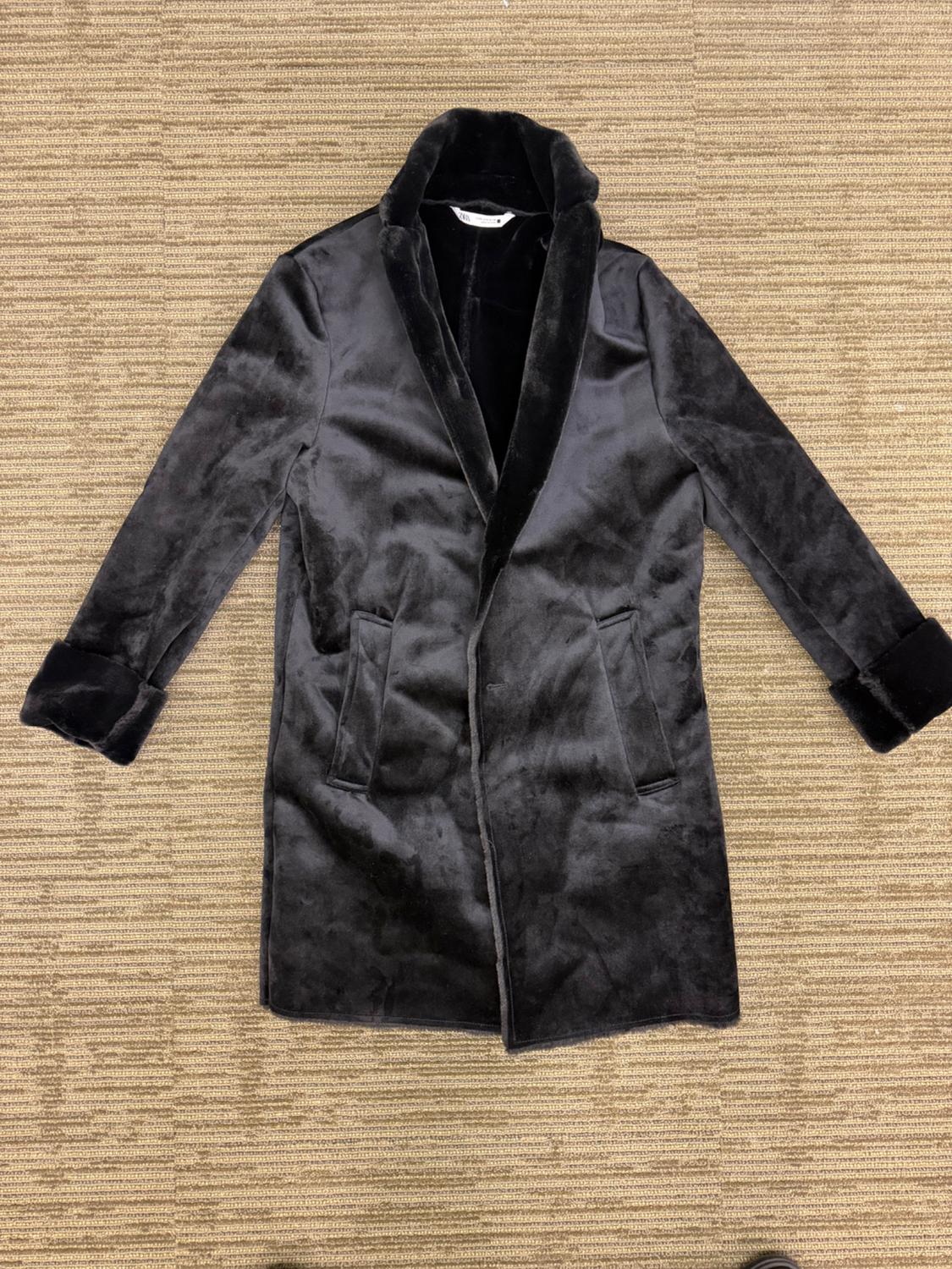 Black coat with a soft-textured finish, with a fur collar and cuffs, laid out over a textured brown surface. Zara brand label on the collar.