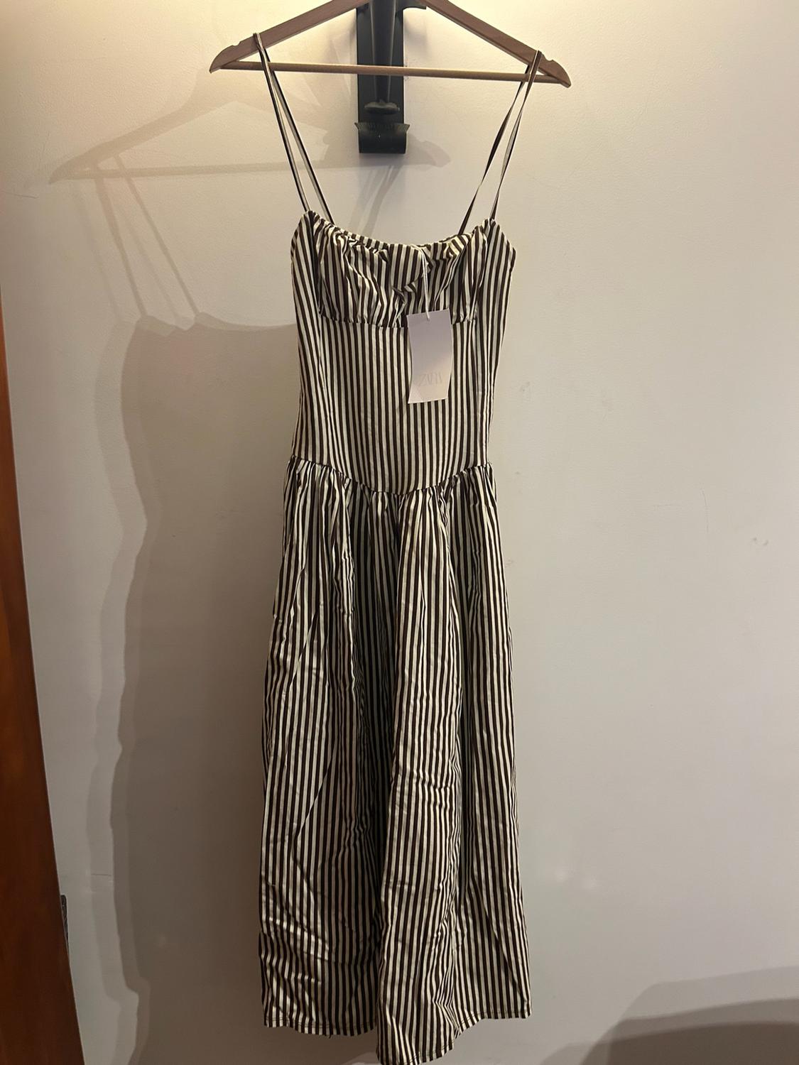 Black-and-white vertically striped spaghetti-strap dress with a ZARA label, hanging on a wooden hanger against a light-coloured wall.