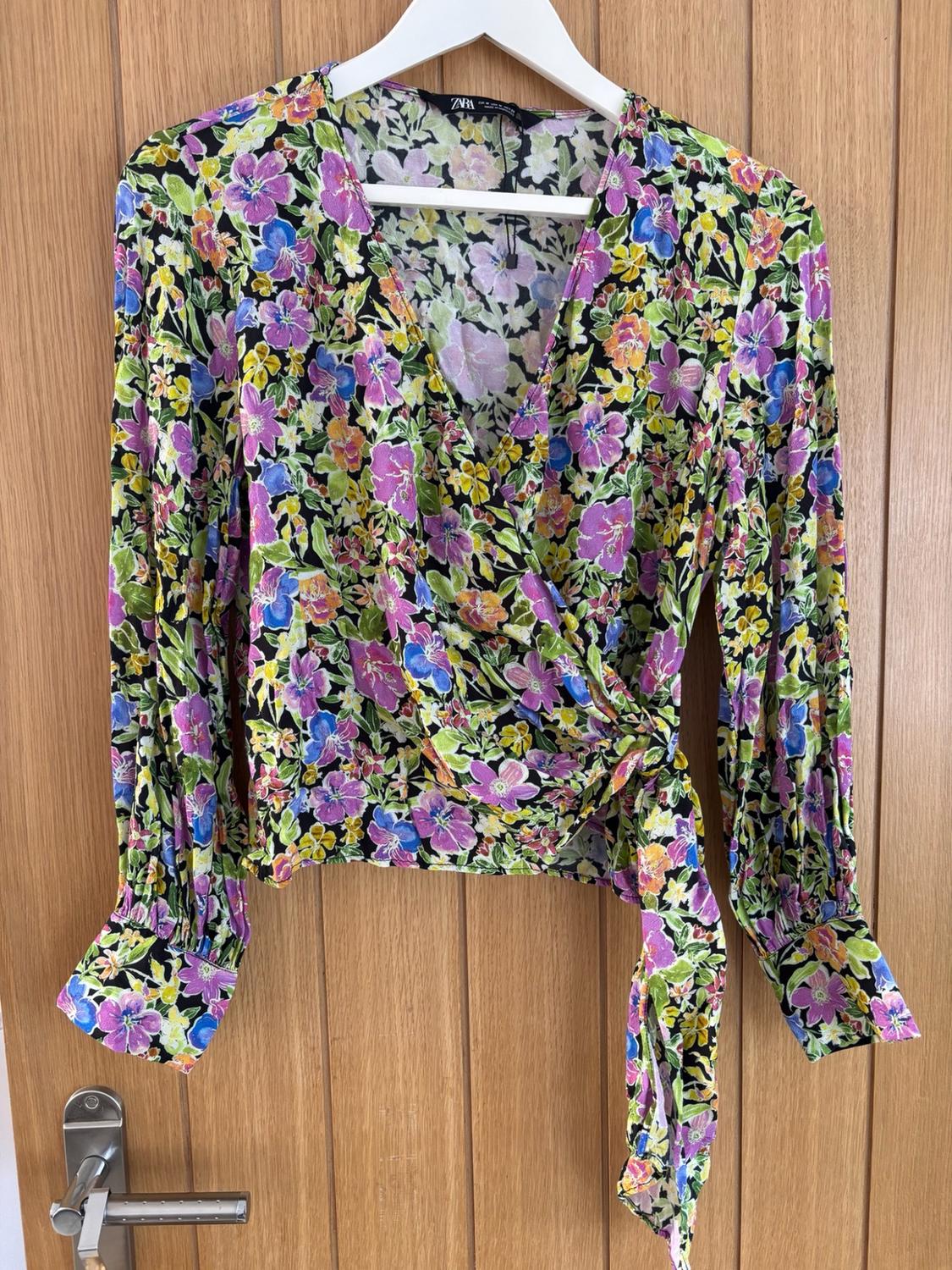 Long-sleeved wrap blouse with floral print and side tie, hanging on a white hanger against a wooden door. Text: ZARA.