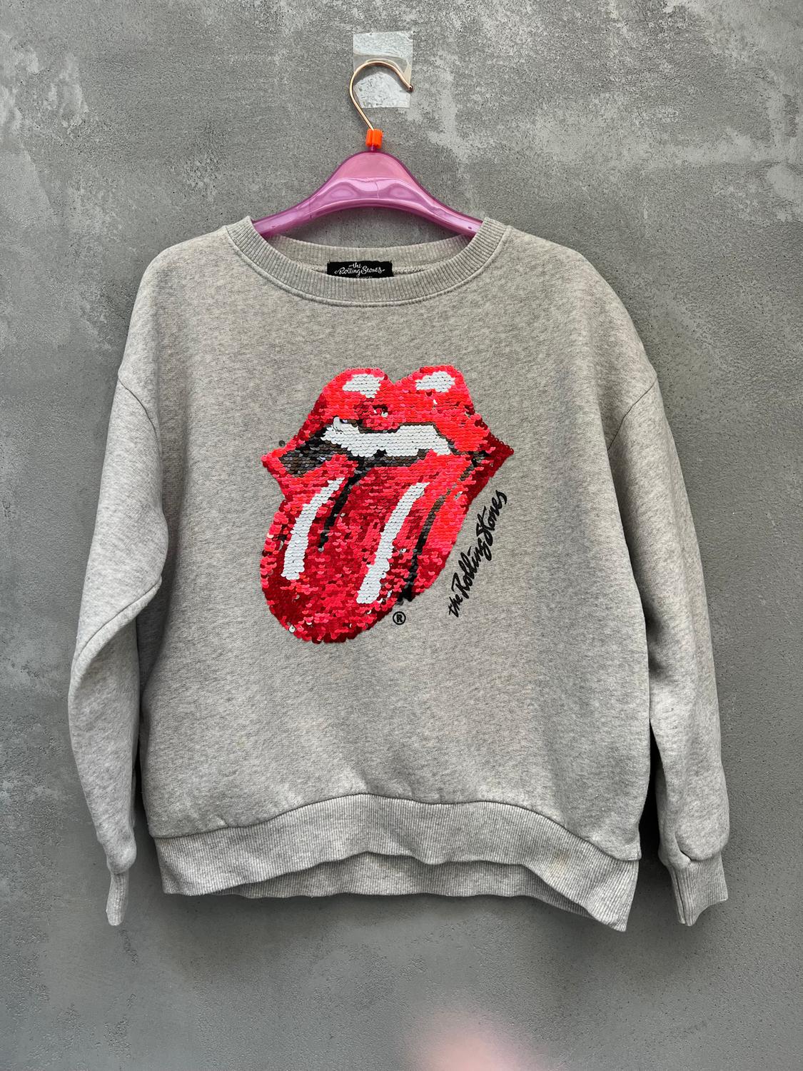 Grey sweatshirt with red tongue logo and sequins, hanging on a purple hanger against a grey wall background. Text: "The Rolling Stones".