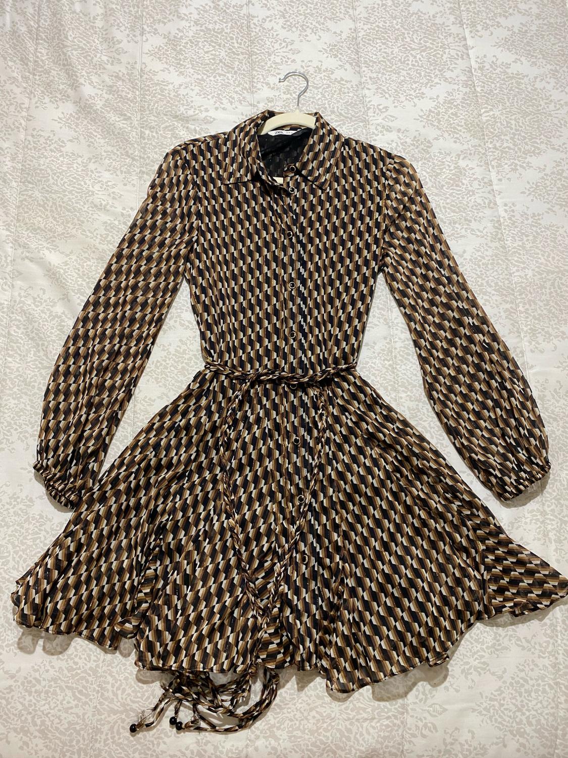 Long-sleeved dress with a geometric print in brown, black, and beige tones, hanging on a hanger against a light background.