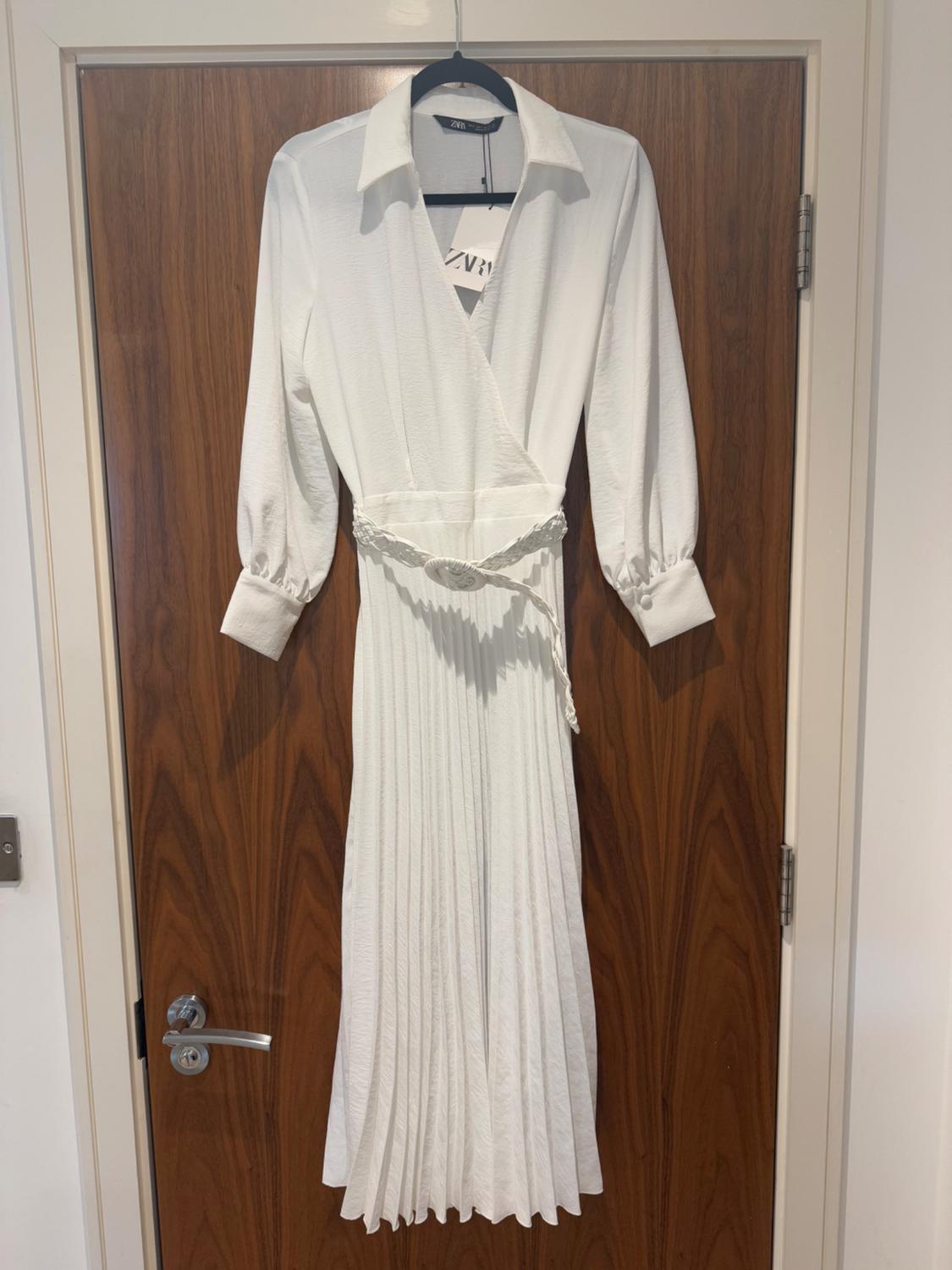 White pleated dress with long sleeves and a braided belt, hanging on a wooden door. Visible label with text "ZARA".