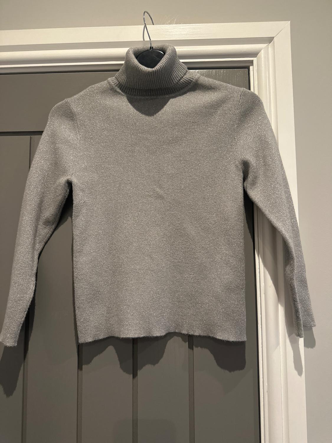 Grey roll-neck jumper hanging on a hanger over a grey door. The texture of the fabric is visible.
