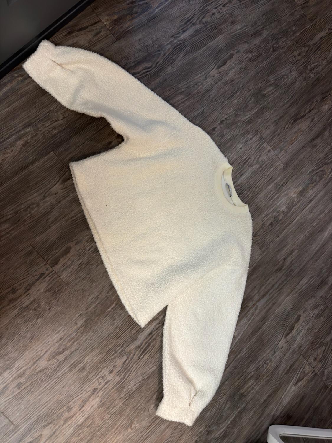Cream-colored sweater with a soft, fluffy texture, a crew neck, and long sleeves, laid out on a wooden floor.