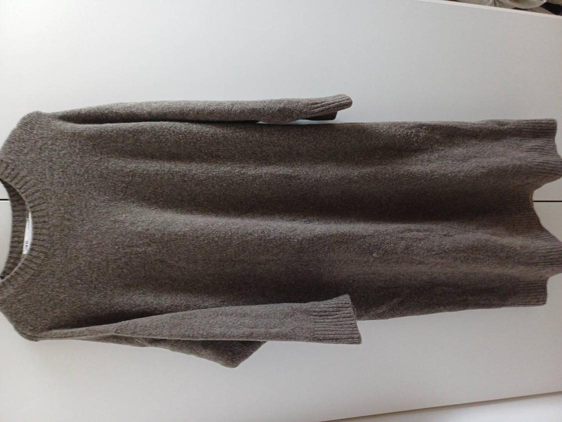 Grey knitted jumper hanging against a white wall. Simple design with a round neck and long sleeves.