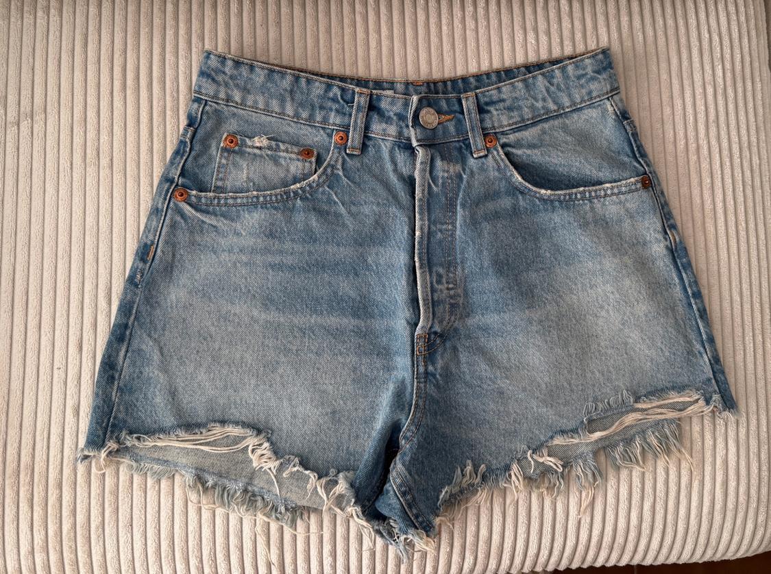 Light blue denim shorts with frayed hem and pockets, on a light ribbed surface. Casual and relaxed style.
