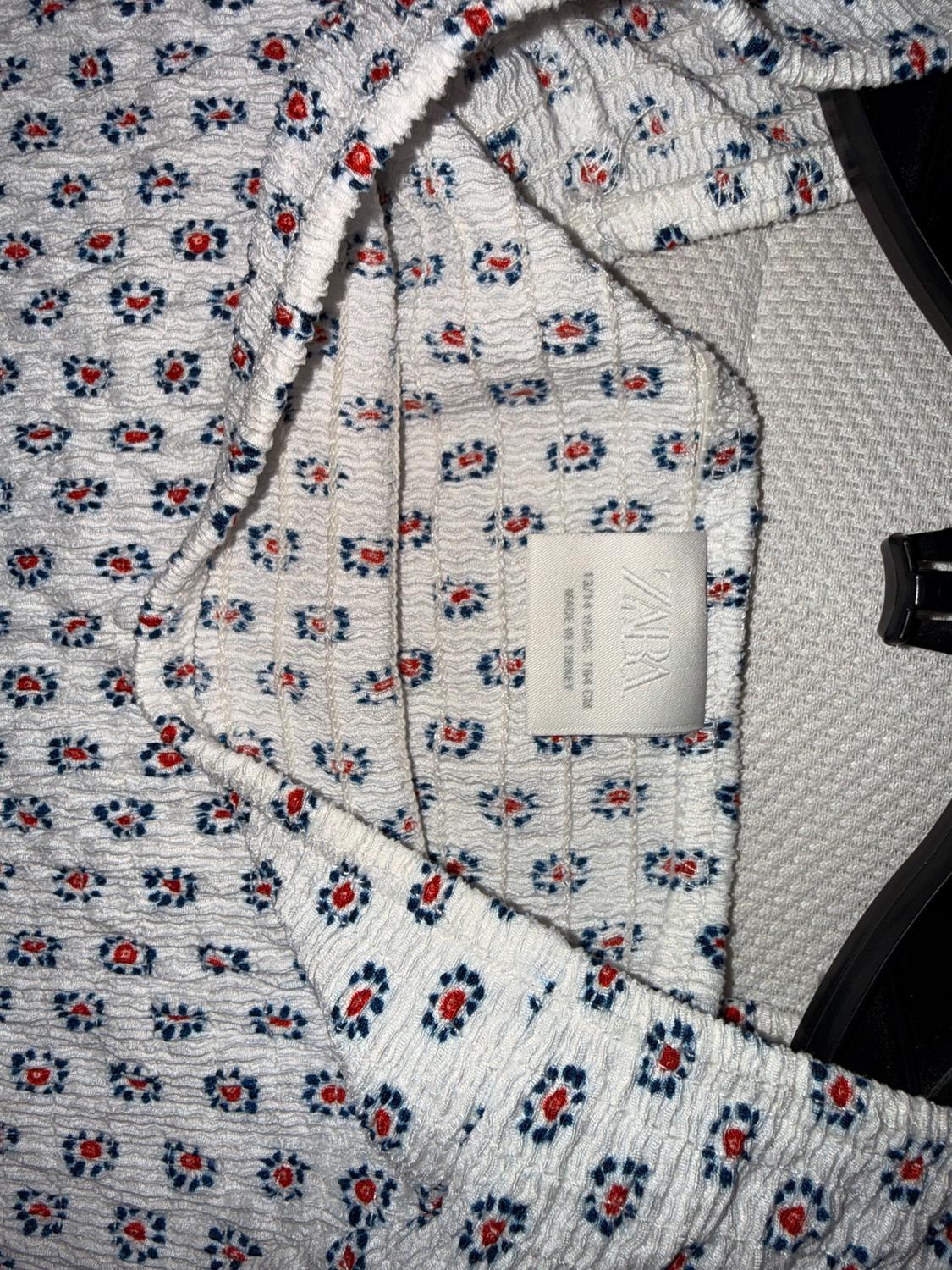 White fabric with blue and red floral print, visible label with "ZARA" text, wrinkled texture, in a soft lighting environment.