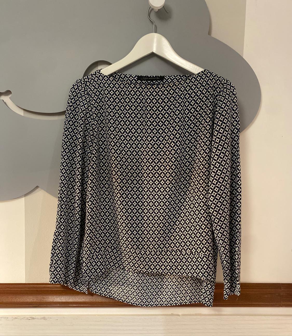 Black and white long-sleeved blouse with a geometric print, hanging on a white hanger against a grey background.