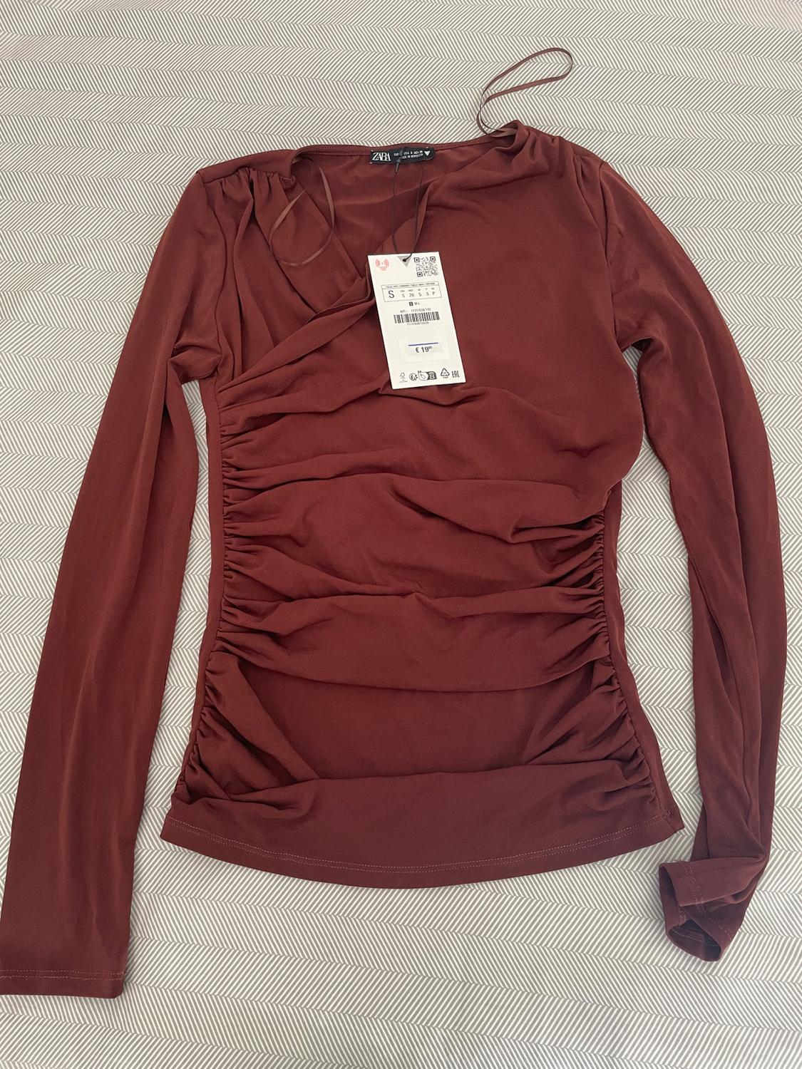 Brown long-sleeved blouse with ruching, visible label with price of €19.95, on a herringbone fabric background.