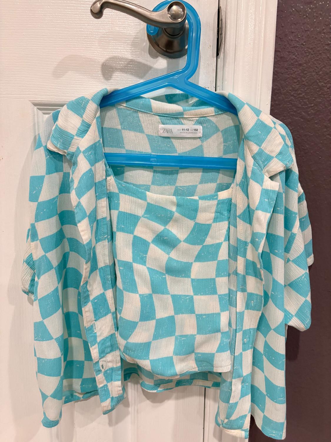 Blue and white shirt with a wavy checkered pattern, hanging on a blue hanger over a white door. Visible tag: "ZARA".