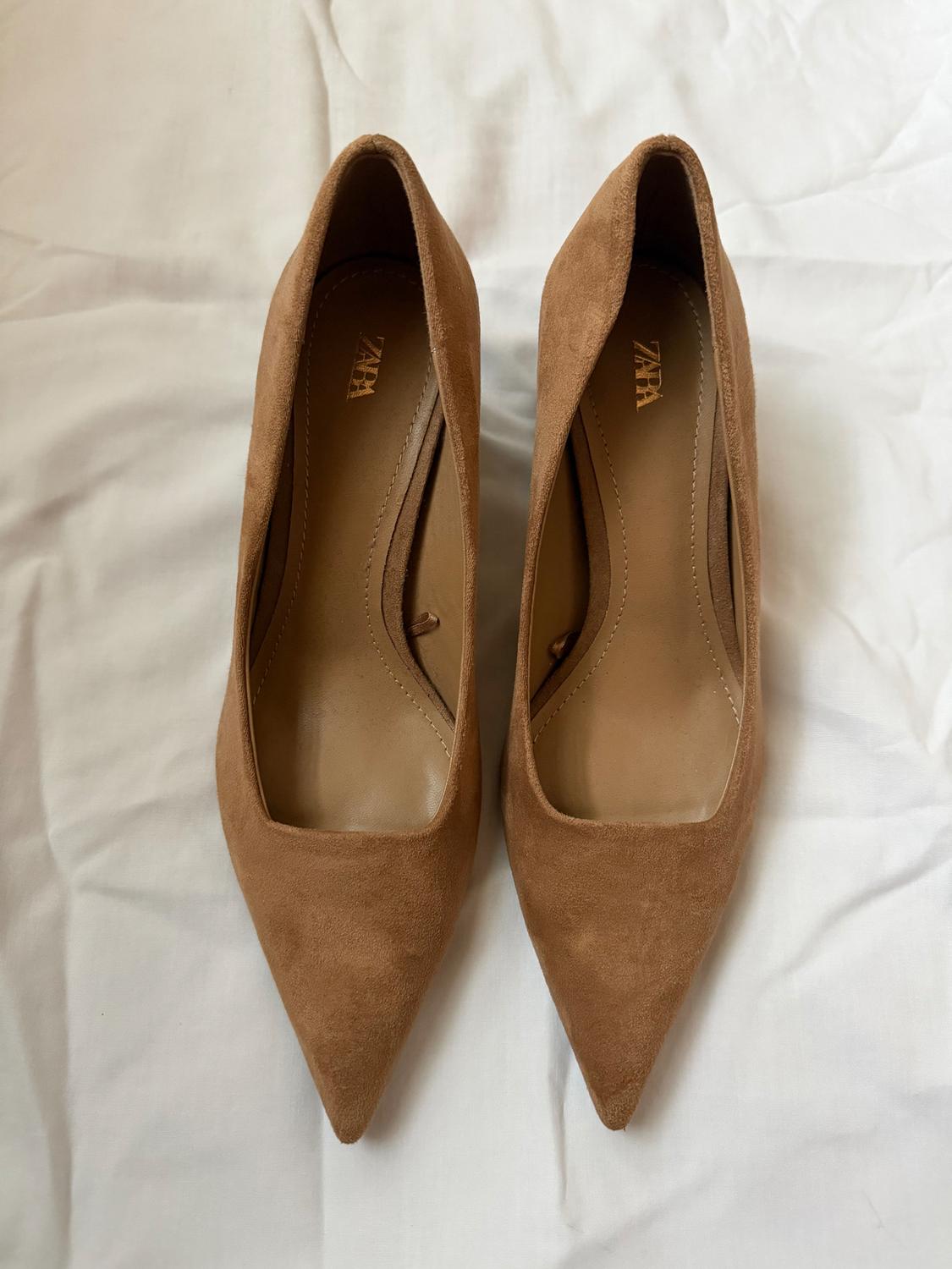 Light brown high heels with pointed toes on white fabric. The brand "Zara" is visible inside the shoes.