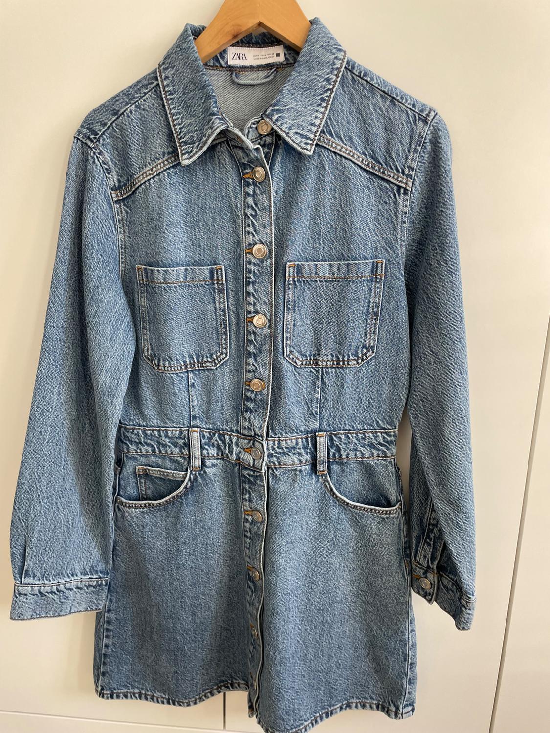 Light blue denim dress with front buttons and pockets, hanging on a wooden hanger. Visible label: "ZARA".