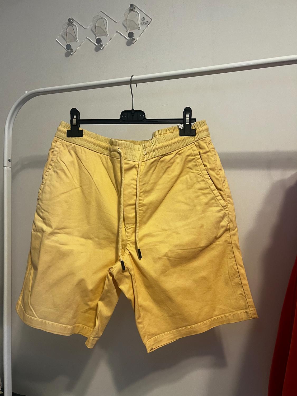 Yellow shorts hanging on a black hanger against a white background. Transparent wall hook visible above.