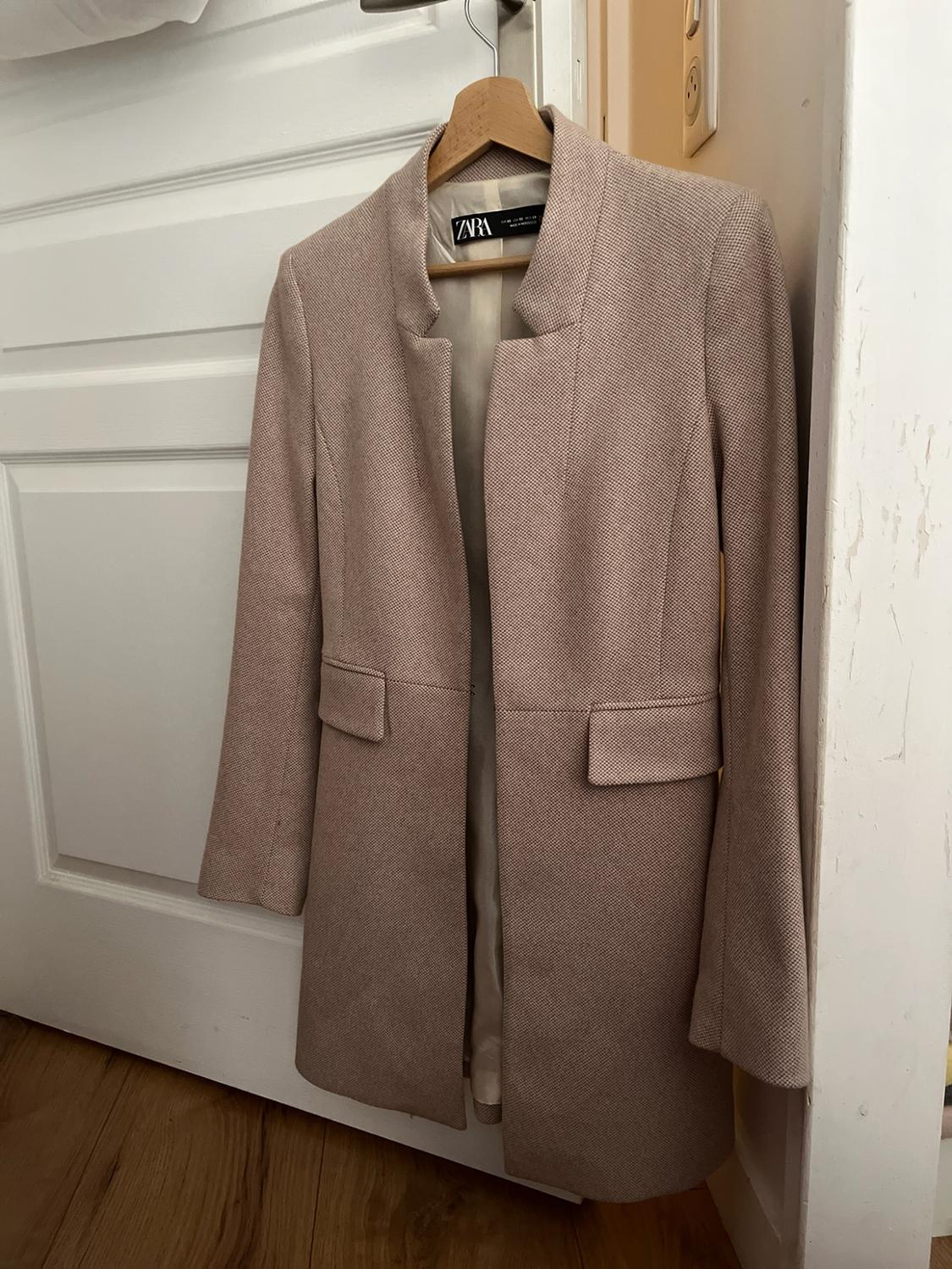 Long beige coat with pockets hanging on a wooden hanger against a white door. Label with the text ZARA. Relaxed style.