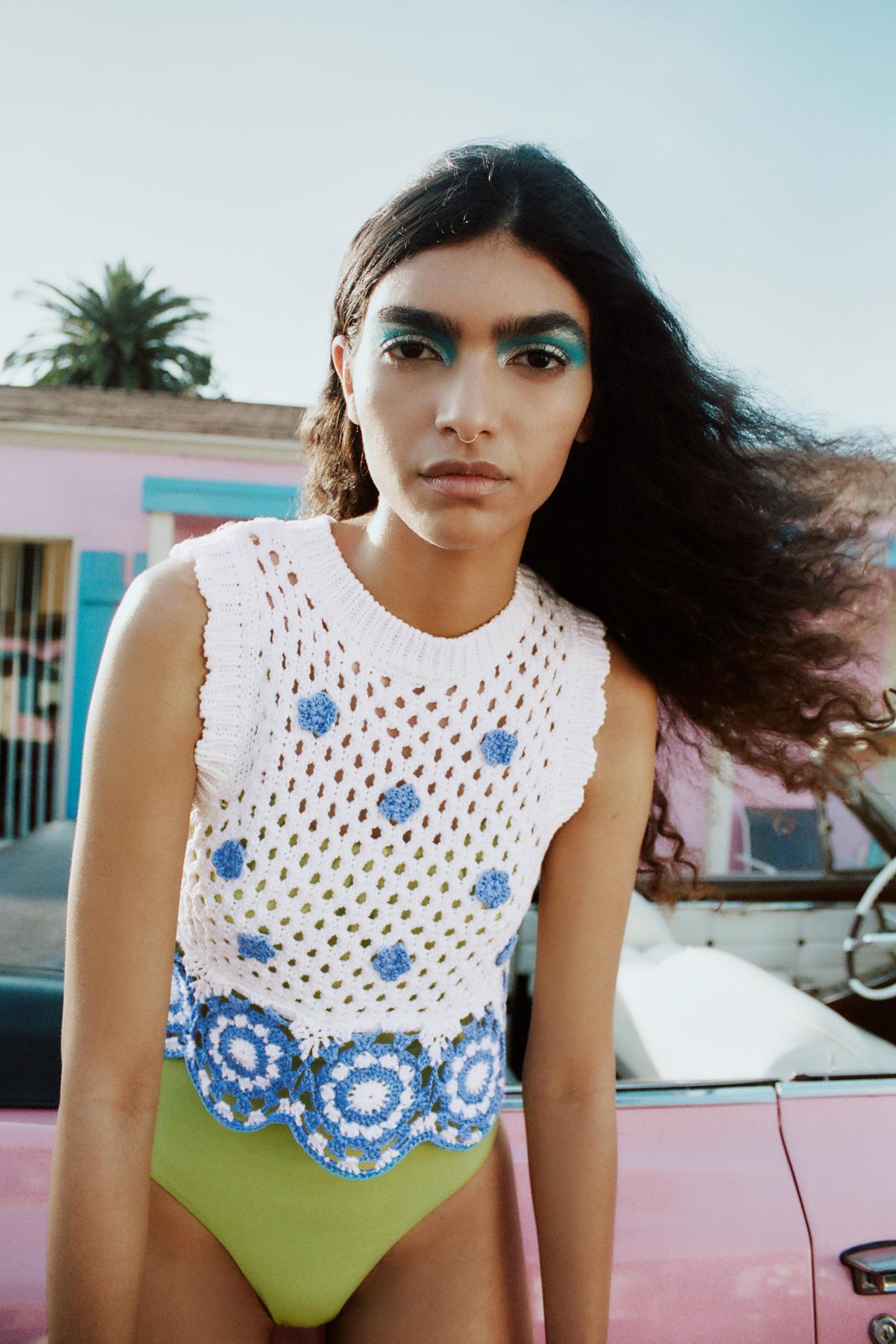 Sleeveless white knit top with ribbed round neck and scalloped edges on the sleeves. Blue crochet mini flower detail on the front and blue and white crochet detailing at the cropped hem, creating a scalloped finish.